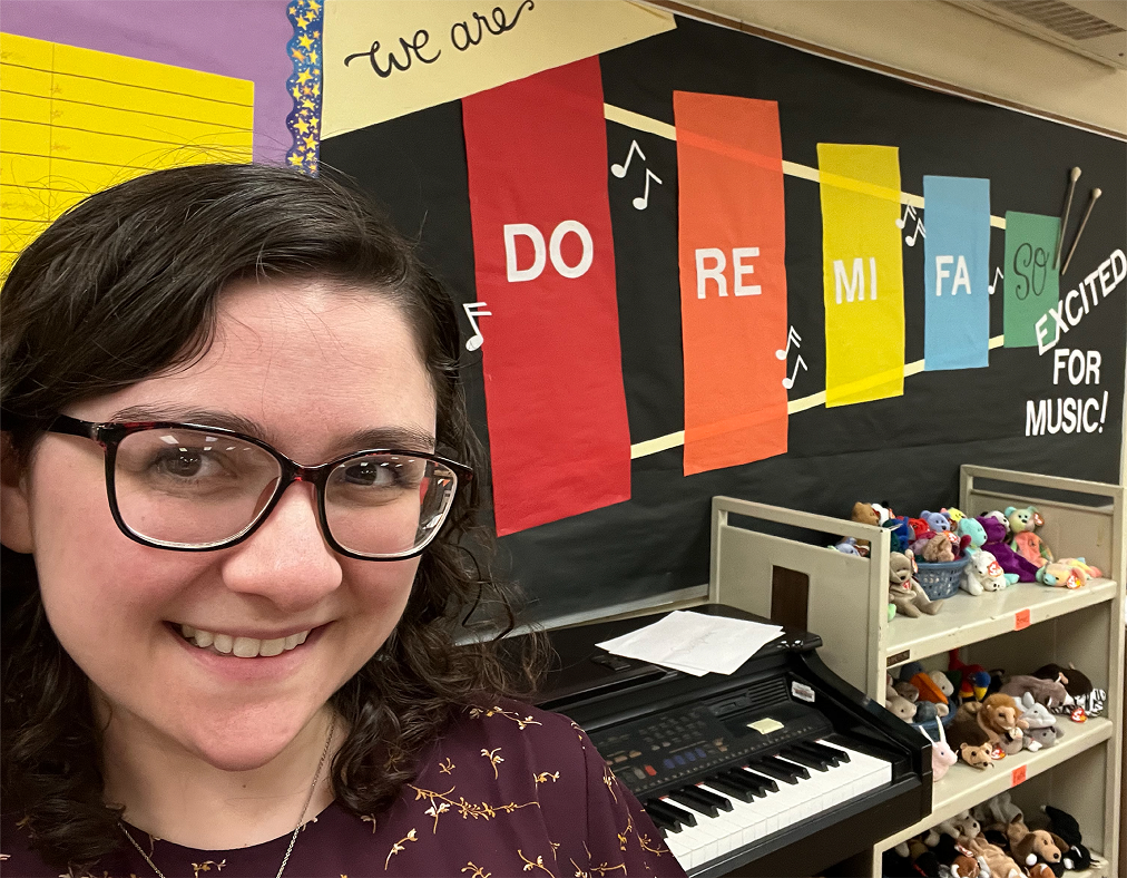 Master of Music in Music Education student, Rebecca Dangerfield, smiling in her classroom in front of a display that says "We are Do Re Me Fa So Excited for Music!"