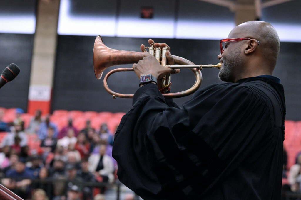 Dana School of Music alum and world-renowned jazz musician, Sean Jones, playing his trumpet at YSU's commencement after giving the commencement speech to our graduates. He's playing while wearing regalia, and there is an audience visible in the background watching from the bleachers.