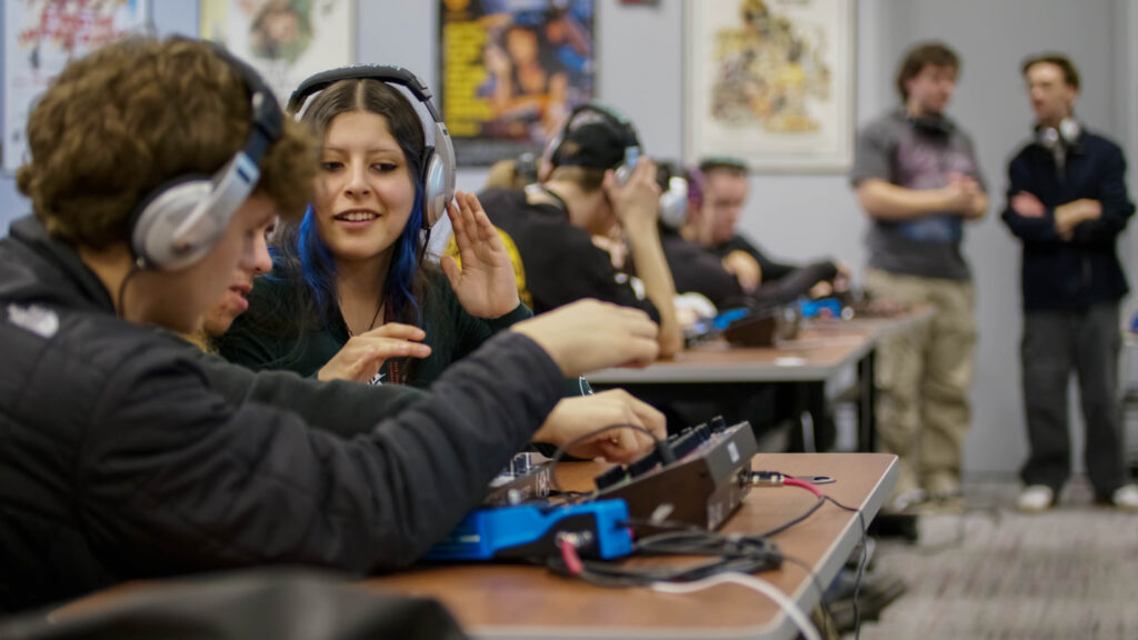 High school students during a workshop learning how to use digital audio workstations. They are in a classroom in YSU's Bliss Hall, wearing headphones and interacting with one another.