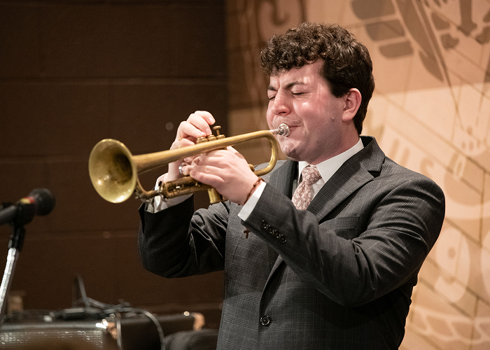 Student playing trumpet during a jazz performance. He is on stage, with a graphic of the YSU official seal behind him.